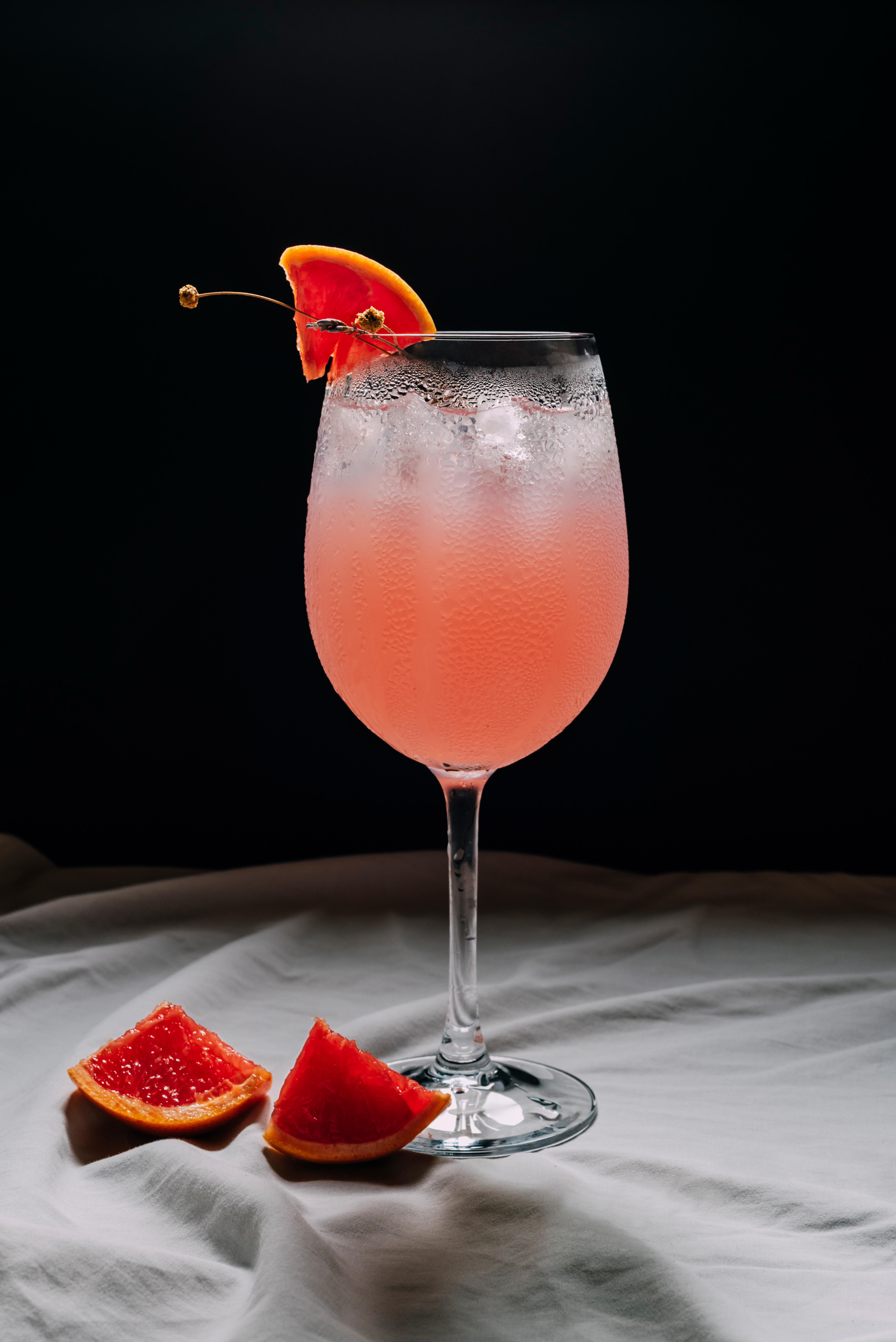 Ein Glas pinkfarbener Grapefruit-Spritz mit Eis und Garnitur auf weißem Stoff vor schwarzem Hintergrund.
